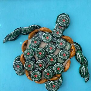 以下が含まれることがあります： ハイネケンのボトルキャップで作られた手作りのカメのアートワーク。カメの体は、明るい青色の背景に配置された、多数の緑と金のボトルキャップで構成されています。ハイネケンのロゴが見えます。