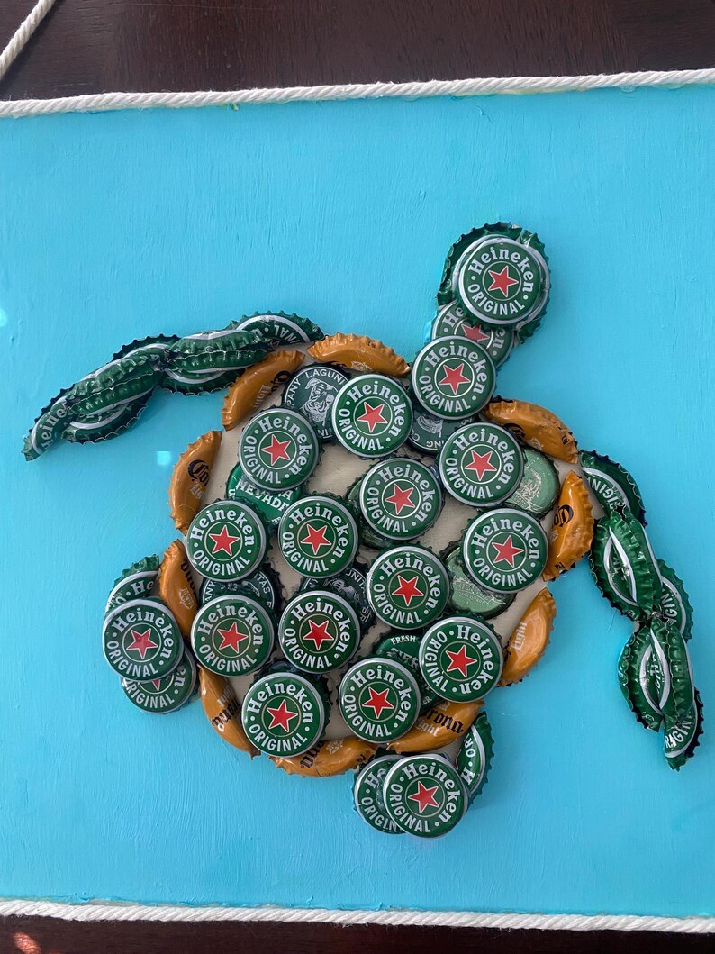 以下が含まれることがあります： ハイネケンのボトルキャップで作られた手作りのカメのアートワーク。カメの体は、明るい青色の背景に配置された、多数の緑と金のボトルキャップで構成されています。ハイネケンのロゴが見えます。