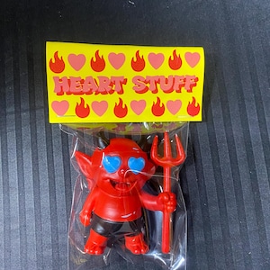 Puede incluir: Una figura de diablo rojo con ojos azules en forma de corazón y un tridente rojo, empaquetada en una bolsa de plástico transparente. Una etiqueta amarilla en la parte superior dice "HEART STUFF" con corazones y llamas rosas.