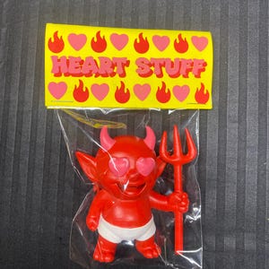 May include: A red devil figurine with heart-shaped eyes and a red trident, packaged in a clear plastic bag. The figurine wears white briefs. A yellow card above reads "HEART STUFF" with pink hearts and flames.