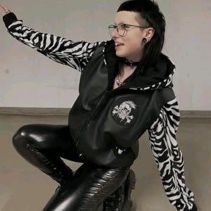 May include: A black leather jacket with zebra print sleeves and a skull patch. The person is wearing black leather pants, a black choker, and round glasses. The jacket has a hood.