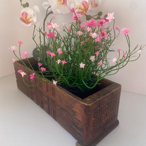 May include: A rectangular wooden planter filled with a mix of artificial flowers. The arrangement includes white orchids with pink and yellow centers, and small pink flowers with green stems. The planter has a dark brown finish.