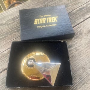 May include: A Star Trek Insignia Collection pin featuring a circular gold-tone base with a central clear stone and a silver-tone triangular overlay. The item is presented in a black box with the text The Official STAR TREK Insignia Collection.