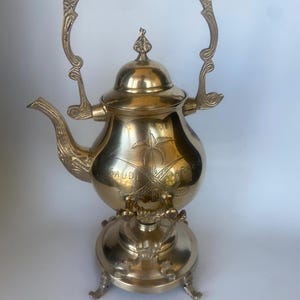 May include: A gold-colored metal teapot with a curved handle and a decorative base. The teapot has a spout and a lid with a small finial. The words "SAUDI ARABIA" are engraved on the body of the teapot, along with a palm tree design.