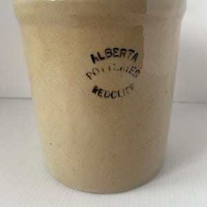 May include: A cylindrical beige stoneware crock with a speckled finish and a thick rim. The front features a black stamped logo that reads ALBERTA POTTERIES REDCLIFF in three lines. The vessel has a smooth, glazed surface and a flat base.