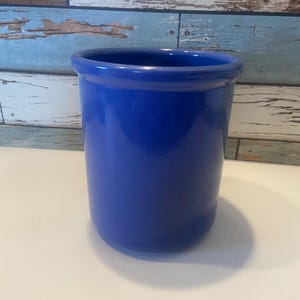 May include: A vibrant cobalt blue ceramic utensil holder. The cylindrical container has a glossy finish and a slightly flared rim. The holder is empty and sits on a white surface, with a weathered wood background.