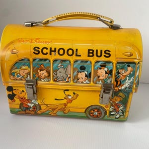 May include: Vintage yellow metal lunchbox featuring a school bus design with Disney characters. The lunchbox has a yellow handle, metal clasps, and illustrations of Mickey Mouse, Pluto, and other characters. The words "Walt Disney" and "SCHOOL BUS" are printed on the lunchbox.