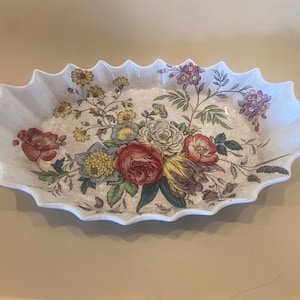 May include: An oval ceramic serving dish with a scalloped edge. The dish is decorated with a floral pattern featuring red, yellow, and purple flowers with green leaves. The background is cream-coloured.