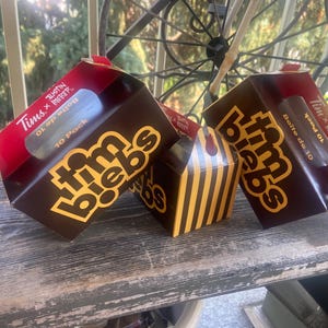 May include: Three brown and yellow cardboard boxes with the text Tim Biebs and Tims x Justin Bieber. The boxes feature a 10 Pack Boite de 10 label. Two boxes are angled, and one smaller box in the centre has vertical yellow and brown stripes.