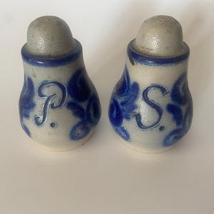 May include: Two ceramic salt and pepper shakers. They are off-white with blue floral patterns and a blue rim. The letters "P" and "S" are visible on the front. The tops of the shakers are light grey in colour.