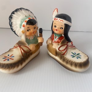 May include: Two ceramic figurines of Native American children in moccasins. One wears a feathered headdress, the other a headband. Both are dressed in traditional clothing and hold small objects. The moccasins are decorated with floral patterns.