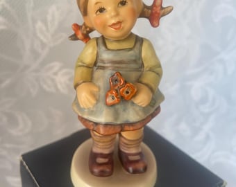May include: A Hummel figurine of a young girl with blonde pigtails, wearing a yellow long-sleeved shirt and a blue-gray dress. She holds three orange flowers. The figurine is on a beige base and stands on a black box.