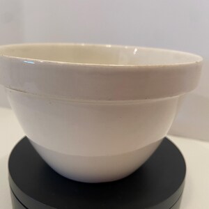May include: A white ceramic mixing bowl with a rounded body and a wide rim. The bowl is sitting on a black surface. The bowl is a classic kitchenware item.