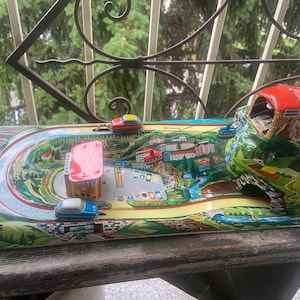 May include: A vintage tin toy playset featuring a colorful lithographed landscape with a winding road, a gas station, and a mountain tunnel. The set includes small metal cars and a cable car mechanism, all set on a flat, rectangular metal base.