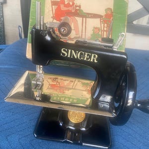 May include: A vintage black Singer toy sewing machine with a hand crank, a silver metal base, and gold lettering. The machine features a circular gold emblem on the front and is positioned in front of its original cardboard box with illustrations.