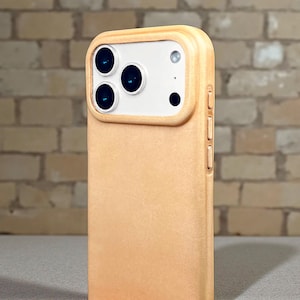 Pode incluir: Uma capa de telefone bege claro com um módulo de câmera com três lentes e um flash. A capa tem um acabamento liso e fosco e cobre as laterais e a parte traseira do telefone. O fundo é uma parede de tijolos.