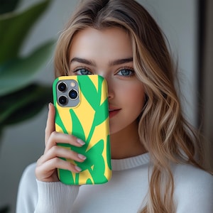 May include: A smartphone case featuring a vibrant design of green leaf-like shapes against a yellow background. The case is held up in front of a person's face, showcasing the phone's camera and overall design. The case is a protective accessory for a smartphone.