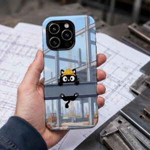 Op de afbeelding: Een smartphonehoesje met een cartoon zwarte kat met een gele veiligheidshelm, zittend op een stalen constructiebalk. De achtergrond toont een blauwe lucht en het stalen frame van een gebouw in aanbouw in een stedelijke omgeving.