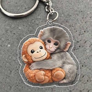 May include: A clear acrylic keychain featuring a cartoon illustration of two hugging monkeys. One monkey is gray, and the other is orange. The keychain has a silver metal ring and chain.