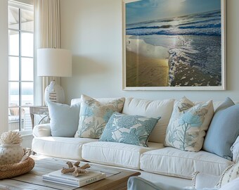 Puede incluir: Un salón con un sofá blanco adornado con cojines azul claro y con estampado floral. Una gran fotografía enmarcada de una escena de playa cuelga de la pared. Una mesa de centro de madera exhibe objetos decorativos, creando un tema costero.