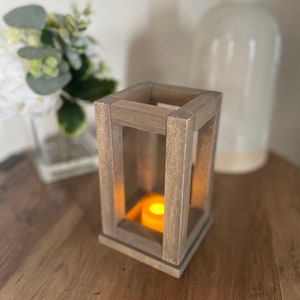 May include: A wooden candle lantern with a warm, glowing candle inside. The lantern is made of natural wood and has a rustic, farmhouse style.
