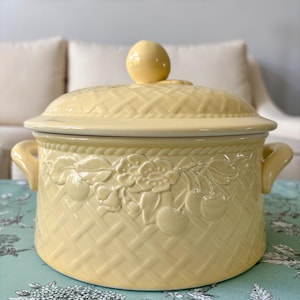 May include: A pale yellow ceramic casserole dish with a matching lid featuring a knob handle. The exterior has a raised basket weave texture and a decorative embossed pattern of cherries and leaves around the upper rim, with two side handles.
