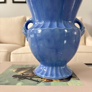 May include: A periwinkle blue ceramic vase with a glossy finish, featuring a wide flared rim, a bulbous body, and two small side handles. The base and the upper section of the vase are decorated with vertical fluted ridges, creating a textured look.