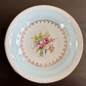 May include: A vintage porcelain bowl with a light blue rim and a white center. The center features a floral bouquet with pink, purple, and blue flowers. Gold trim accents the rim and the floral design. The bowl is likely for serving.