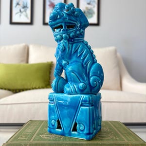 May include: A vibrant turquoise ceramic Foo Dog statue, perched atop a decorative base with cut-out designs. The statue is placed on a vintage green book, with a neutral background of a sofa and framed artwork.
