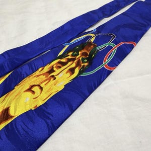 May include: A blue silk tie with a yellow and brown dragon design and green details. The tie also features the Olympic rings in red, yellow, green, and blue. The tie is made of a shiny, smooth fabric.