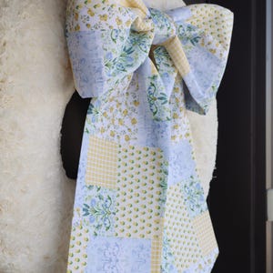 May include: A white, fluffy wreath with a large bow made of patchwork fabric. The bow features patterns of yellow gingham, blue floral designs, and small yellow flowers. The wreath is hanging on a dark door.