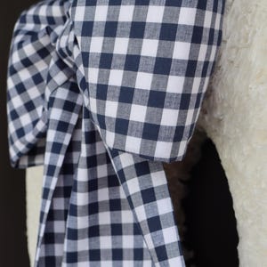 May include: A large bow made of navy blue and white gingham fabric. The bow is attached to a white, textured surface. The fabric is folded and draped to create the bow shape, with the gingham pattern clearly visible.