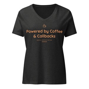 May include: A dark charcoal grey V-neck t-shirt featuring a small coffee cup icon and the orange text "Powered by Coffee & Callbacks" followed by "MEDICAL ASSISTANT EDITION" with a horizontal line underneath the text on the front of the shirt.