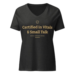 May include: A charcoal grey V-neck t-shirt featuring a gold graphic print. The text reads "Certified in Vitals & Small Talk" with "MEDICAL ASSISTANT EDITION" below it, accompanied by a small shield icon with a checkmark centred above the text.