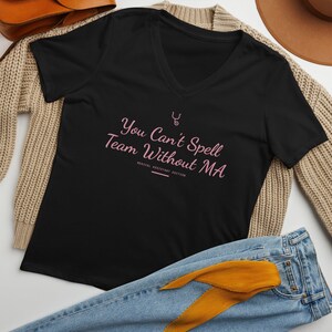 May include: A black V-neck t-shirt featuring light pink text that reads You Can't Spell Team Without MA, with a small stethoscope icon above the text and the words MEDICAL ASSISTANT EDITION printed in smaller letters below the main phrase.