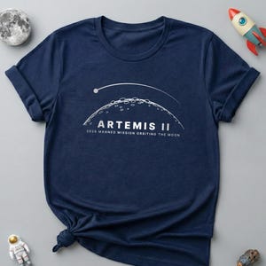May include: Navy blue t-shirt with the text "ARTEMIS II 2026 MANNED MISSION ORBITING THE MOON" in white. The design includes a stylized depiction of the moon and a trajectory line. A toy rocket, astronaut, and moon are also visible.