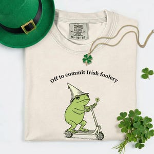 May include: A cream-colored t-shirt with the text "Off to commit Irish foolery" and a cartoon frog riding a scooter. A green hat with a gold buckle and a shamrock necklace are also visible. A bundle of shamrocks is on the right.