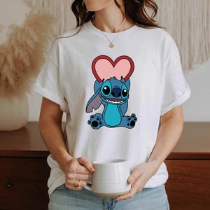 May include: White t-shirt featuring a cartoon character holding a pink heart. The character is blue with large ears and a friendly expression. The shirt is a casual style, suitable for everyday wear.