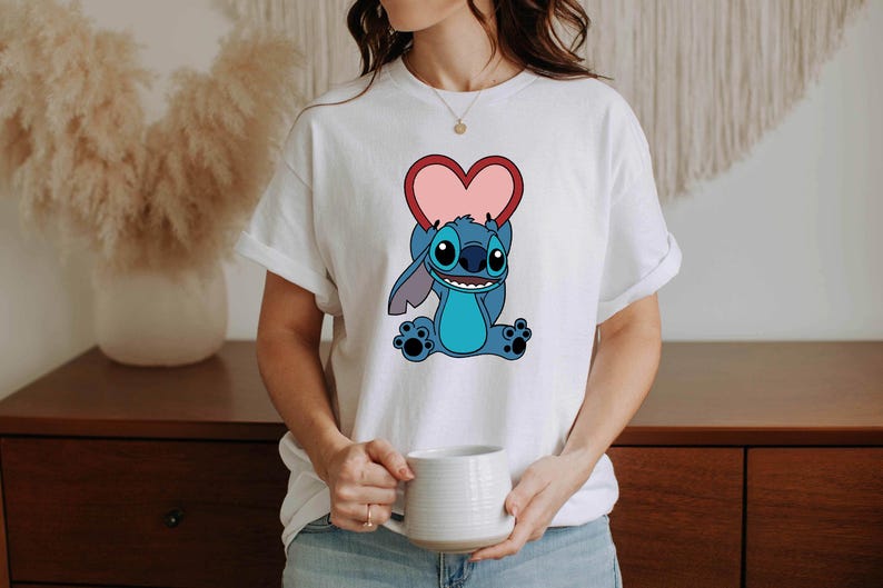 May include: White t-shirt featuring a cartoon character holding a pink heart. The character is blue with large ears and a friendly expression. The shirt is a casual style, suitable for everyday wear.