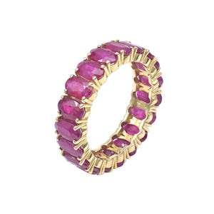 May include: An eternity band ring crafted from yellow gold, featuring a continuous row of oval-cut, deep pinkish-red rubies set in individual prongs that encircle the entire circumference of the band, creating a uniform and vibrant gemstone display.