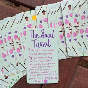 May include: A stack of rectangular tarot cards with a light green background featuring a pattern of acorns, flowers, shells, and purple hearts. The top card displays purple handwritten text: The Snail Tarot, followed by a thank you note from Rowan.