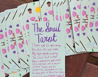 May include: A stack of rectangular tarot cards with a light green background featuring a pattern of acorns, flowers, shells, and purple hearts. The top card displays purple handwritten text: The Snail Tarot, followed by a thank you note from Rowan.