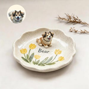 May include: A cream-colored, speckled ceramic dish with a scalloped edge, featuring painted yellow tulips and the name Bear in black text. A small, three-dimensional figurine of a brown and white dog sits in the center of the dish.
