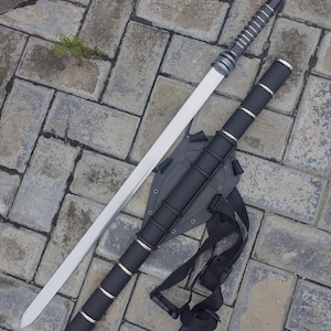 Puede incluir: Una espada plateada y negra con una hoja blanca descansa junto a su vaina negra. La vaina tiene una correa negra y anillos plateados. El mango de la espada es negro con detalles plateados.