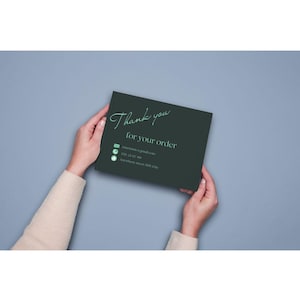 以下が含まれることがあります： 2 つの手で持たれた濃い緑色のカード。筆記体で「Thank you for your order」と書かれています。メールアドレスと電話番号を含む連絡先の詳細は、その下に印刷されています。背景は無地の水色です。