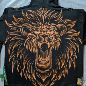 Può includere: Felpa con cappuccio nera con un grande disegno di testa di leone sul retro. La criniera e i lineamenti del leone sono di colore arancione, creando un contrasto sorprendente. La felpa ha un cappuccio con coulisse e una tasca a marsupio.