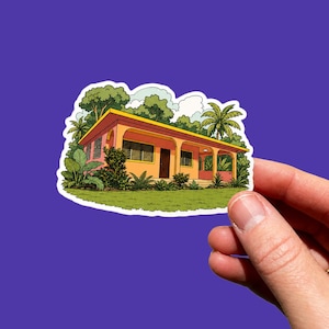 Può includere: Un adesivo fustellato con un'illustrazione colorata di una casa a un piano con pareti color pesca, un bordo del tetto giallo e un portico. La casa è circondata da una rigogliosa vegetazione tropicale, palme e un prato verde.