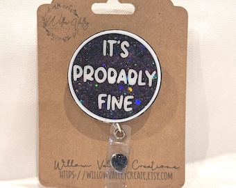 Può includere: Un porta badge con un design rotondo e glitterato che recita "IT'S PRODABLY FINE" in lettere bianche. Il badge è attaccato a una carta marrone con il logo e l'indirizzo del sito web di Willow Valley Creations.