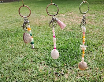 May include: Three keychains with seashell pendants and beaded details suspended from a wooden rod. Each keychain has a unique design, including colourful beads, a pink tassel, and a starfish charm. The keychains are set against a background of green grass.