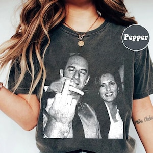 May include: Dark gray t-shirt featuring a black and white image of two people. One person is giving the middle finger. The t-shirt has a distressed look and the word "Pepper" is printed on the upper right.